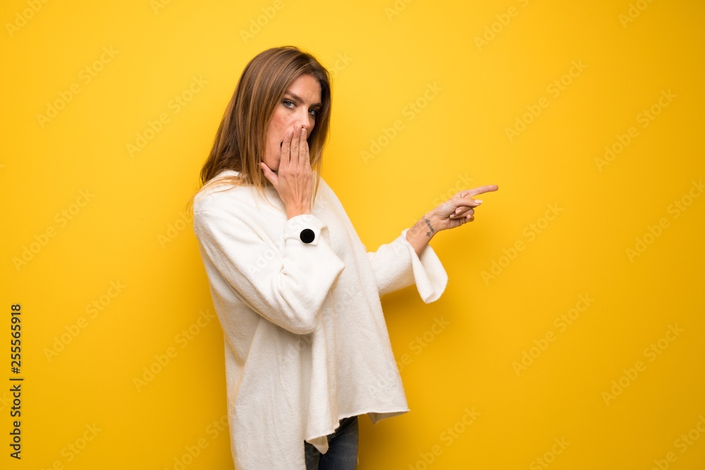 Blonde woman over yellow wall pointing finger to the side with a surprised face Blonde woman over yellow wall pointing finger to the side with a surprised face
