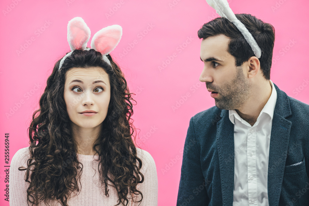 Young happy couple on the pink background. During this, on the head is a rabbit ears. Looking at the camera fool. Doing big eyes. During this man he stares at his wife. Easter. Young happy couple on the pink background. During this, on the head is a rabbit ears. Looking at the camera fool. Doing big eyes. During this man he stares at his wife. Easter.