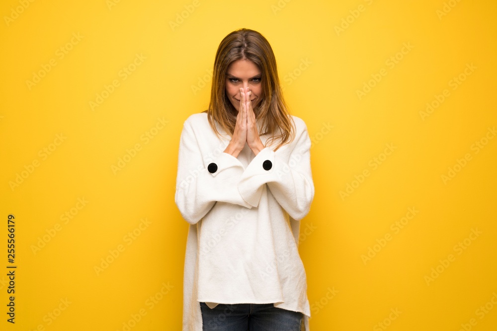 Blonde woman over yellow wall keeps palm together. Person asks for something Blonde woman over yellow wall keeps palm together. Person asks for something