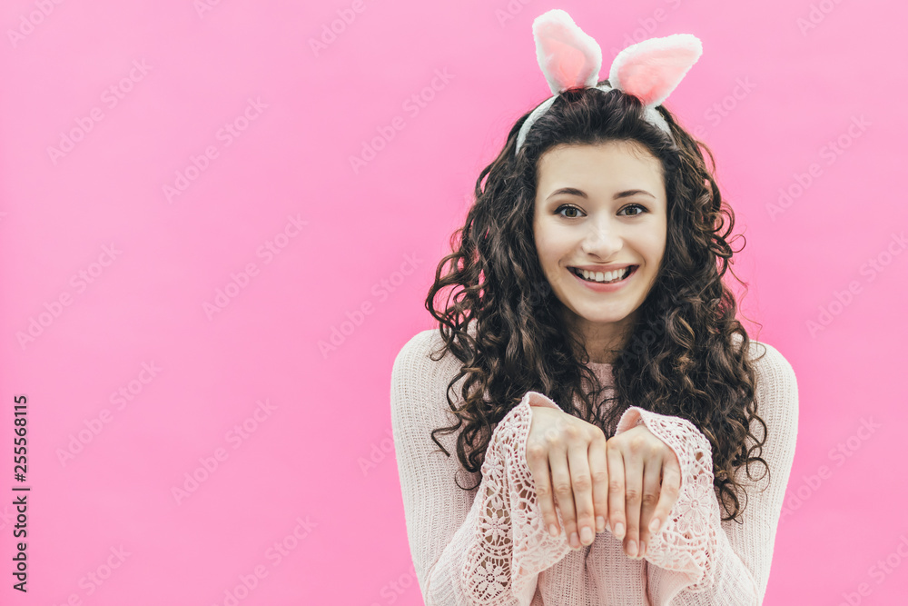 Young pretty girl standing on a pink background. On the head of the ears of the bunny. During this time, he put his hands like a bunny, smiling. Happy Easter Young pretty girl standing on a pink background. On the head of the ears of the bunny. During this time, he put his hands like a bunny, smiling. Happy Easter