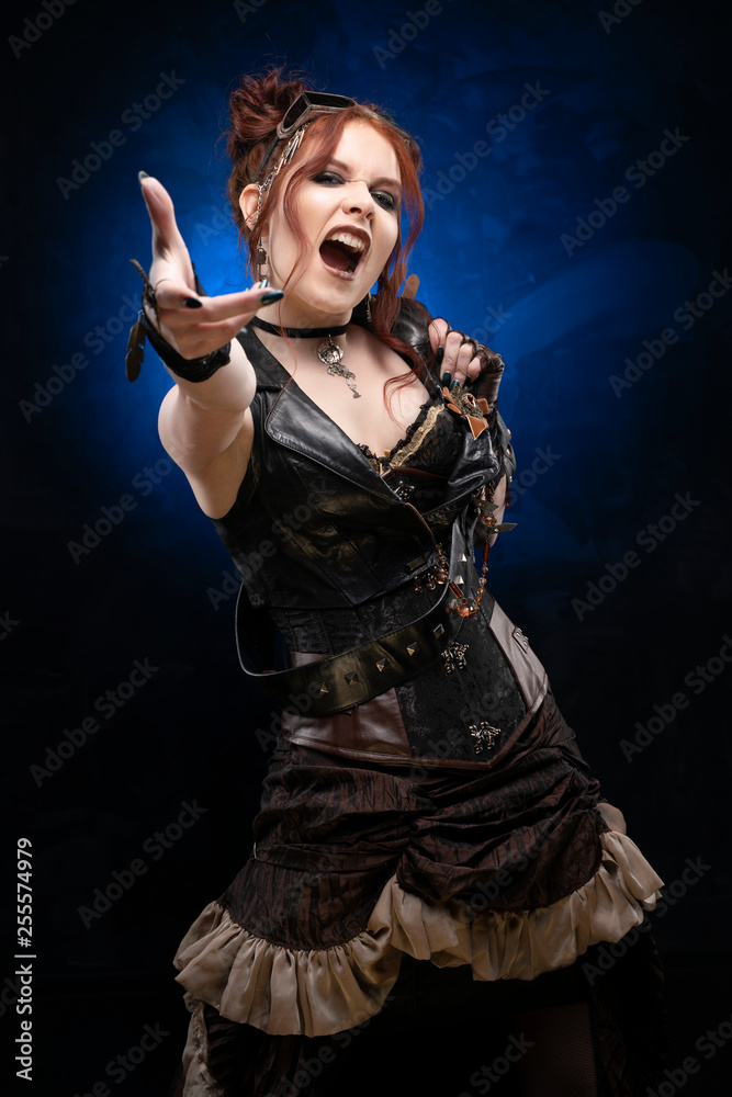 Beautiful redhead cosplay girl wearing a Victorian-style steampunk costume with a big breast in a deep neckline shouts and holds out her hand on a black and blue background
