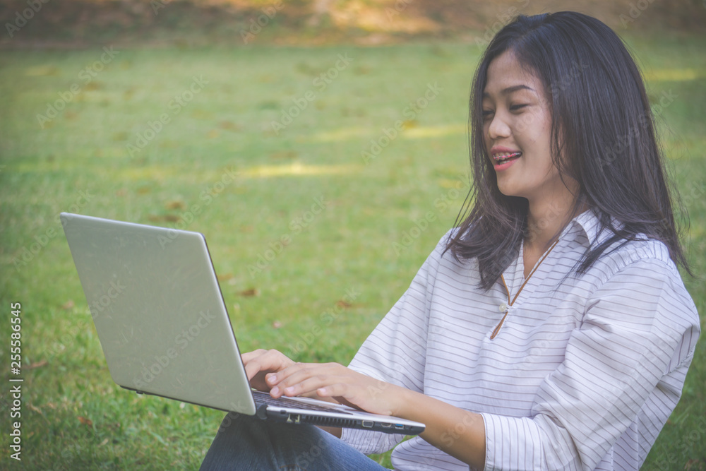 The asia woman is playing computer on a notebook.She sits and play laptop in park.