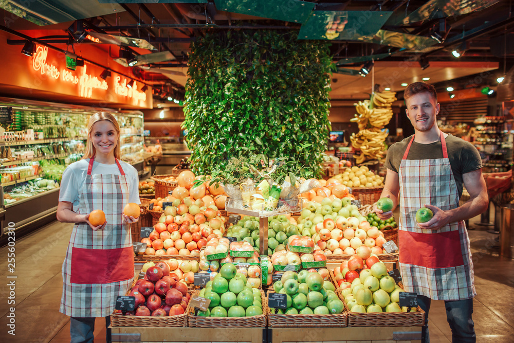 Young man and woman stand at fruit boxes in grocery store. They hold citrus in hands and smile. Workers look straight and pose on camera. Young man and woman stand at fruit boxes in grocery store. They hold citrus in hands and smile. Workers look straight and pose on camera.