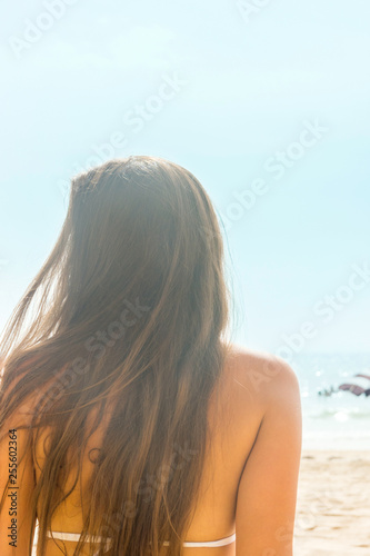Beautiful young caucasian woman girl with long chestnut hair golden tan in white swimming suits sits on beach looking at sea. Bright sunlight blue sky. Summer travel vacation wanderlust concept