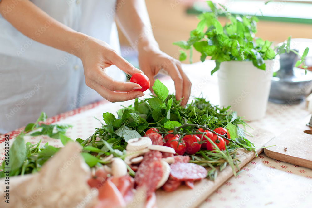 Woman is cooking in home kitchen. Female graceful hands hold cherry tomatoes and greens. Ingredients for preparing italian or french food are on table on wooden boards. Lifestyle moment. Close up. - obrazy, fototapety, plakaty 