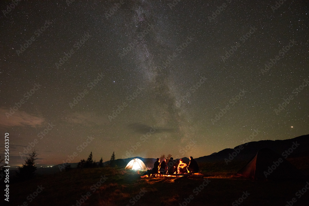 Summer night camping under stars. Group of five people, men and woman with guitar sitting by burning campfire at tourist tents under dark sky with bright sparkling stars and Milky Way constellation.