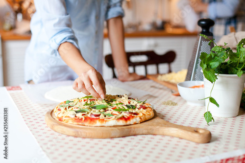 Woman is cooking pizza in cozy home kitchen. Girl is decorating italian dinner with greens, fresh basil, arugula. Homemade pizza is served on wooden board on table. Lifestyle moment. Close up.