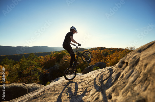 Professional sportsman cyclist jumping on trial bicycle on top of big boulder. Male biker making acrobatic stunt on summer evening, blue sky and sunset on background. Concept of extreme sport photo