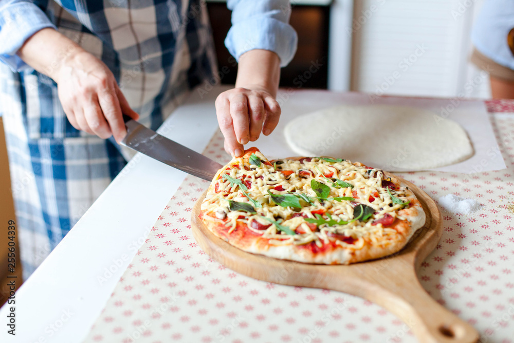 Woman cuts hot pizza on wooden board in cozy home kitchen. Cooking process of italian family dinner at home. Female hands hold knife and slice of pizza. Lifestyle moment.