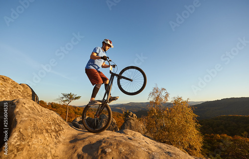 Professional cyclist riding on back wheel on trial bicycle. Sportsman biker balancing on the edge of big boulder on the top of mountain at sunset. Concept of extreme sport active lifestyle