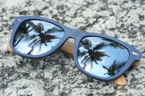 Blue sunglasses with a reflection of the coconut trees on the lens, which is placed on the stone.