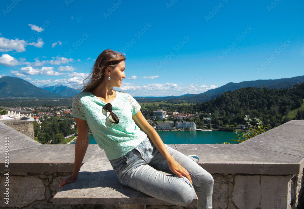 Slovenia, Lake Bled. The view from the Mariinsky temple on the island of the lake on its surroundings. A sunlit girl tourist sits on a stone fence and admires the beautiful scenery.