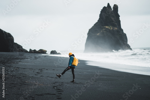 Young hipster girl walks black beach Vík in Iceland, yellow backpack and hat