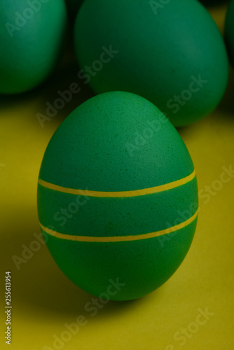 One green painted Easter egg with two yellow stripes stands in front of green eggs on a yellow background. Happy Easter holiday card or banner.