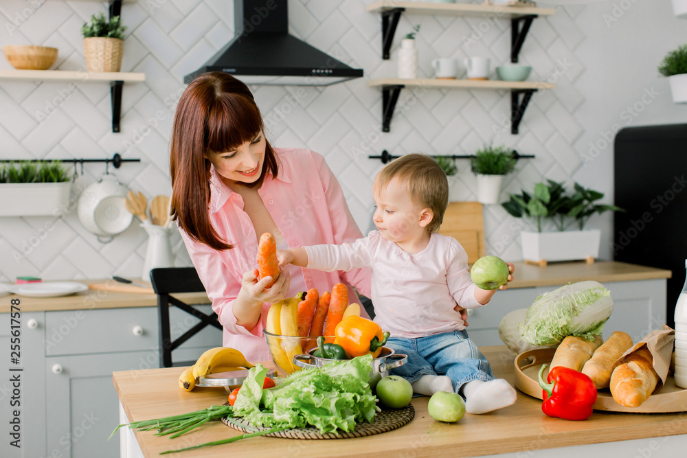 Happy mother showing fresh vegetables to her little baby girl sitting on the wooden table at home kitchen. Family, food, healthy eating, cooking and people concept