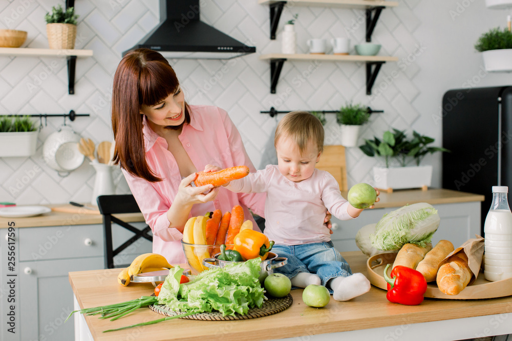 Happy mother showing fresh vegetables to her little baby girl sitting on the wooden table at home kitchen. Family, food, healthy eating, cooking and people concept