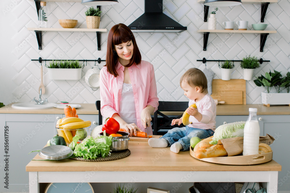 Young pretty mother in rose shirt preparing fresh breakfast and chopping carrot at home kitchen. Little baby girl holding banana and sitting on the wooden table with fresh food, milk and bread