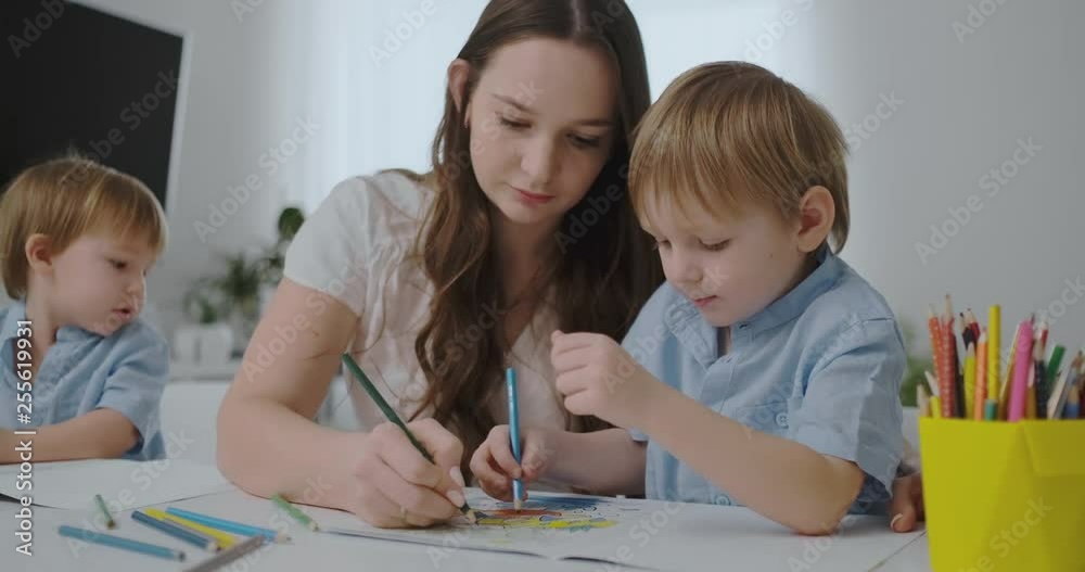 A young mother with two children sitting at a white table draws colored pencils on paper helping to do homework A young mother with two children sitting at a white table draws colored pencils on paper helping to do homework