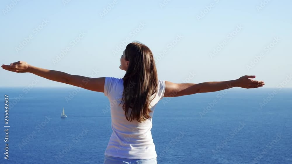 Young woman in white t-shirt raising arms up at cliff against breathtaking view of blue Mediterranean sea. Carefree female outstretching hands. Concept of freedom and inspiration Young woman in white t-shirt raising arms up at cliff against breathtaking view of blue Mediterranean sea. Carefree female outstretching hands. Concept of freedom and inspiration