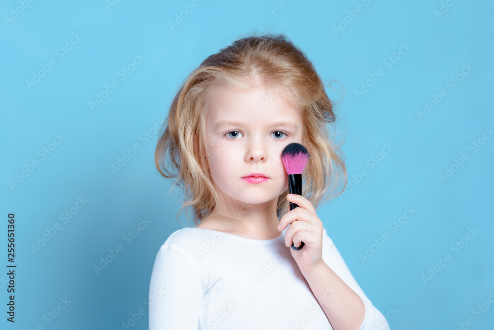 The child put a makeup brush to his cheek. Beautiful girl with blond hair, little model. Bright advertising photography on a blue background. The child put a makeup brush to his cheek. Beautiful girl with blond hair, little model. Bright advertising photography on a blue background.