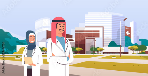 arabic couple man woman doctors in hijab and white uniform standing together in front of hospital building modern medical clinic exterior cityscape background portrait horizontal