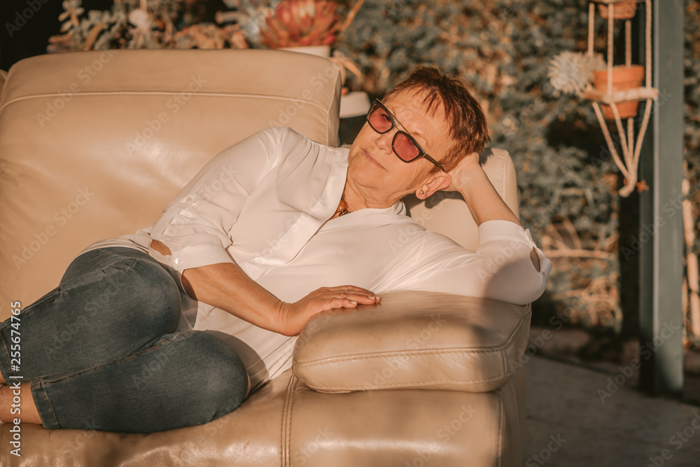 Obraz premium Portrait Of A Cool Senior Woman With Red Short Hair And Trendy Sunglasse Wearing Denim And Blouse. Portrait of an old woman relaxing on a sofa in the garden with a warm light of sunset.