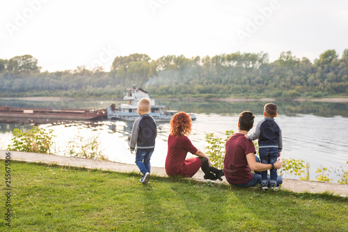 Parenthood, childhood and nature concept - Family sitting on the green ground and looking at small boat