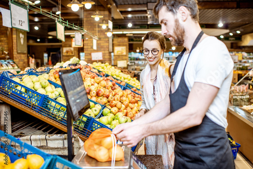 Handsome worker in uniform helping young woman customer to weigh apples on the scales in the supermarket photo