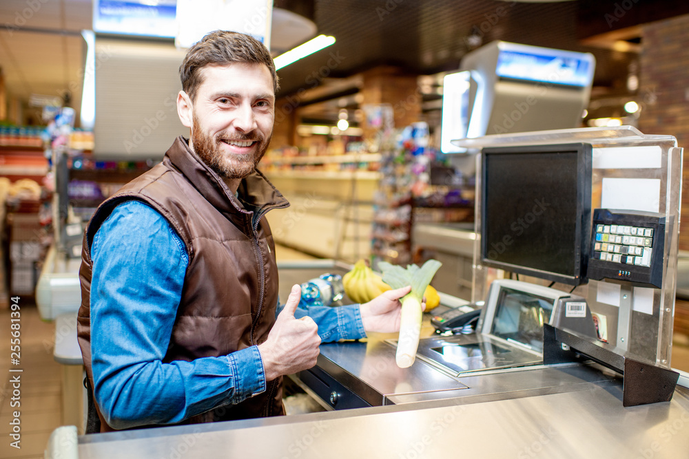 Portrait of a happy and cheerful man as a cashier, sitting at the cash register in the supermarket Portrait of a happy and cheerful man as a cashier, sitting at the cash register in the supermarket