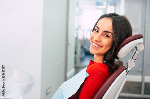 New life with new teeth.  Gorgeous girl wearing red sweater in the stomatology room full of day-light and white colors is smiling with her new  white eye-catching smile.
