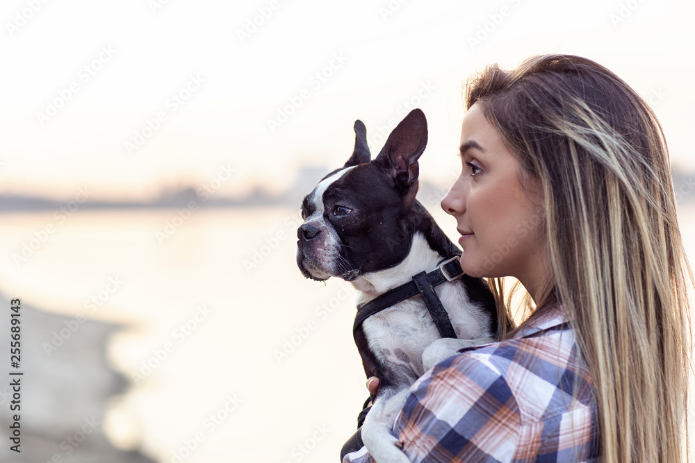 Beautiful girl holding on her hands boston terrier black and white dog on sandy beach near the river