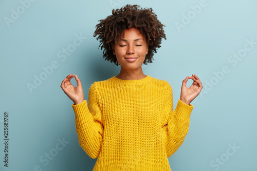 Satisfied dark skinned young woman makes okay gesture with both hands, keeps eyes closed, dressed in yellow clothes, practices yoga after work, isolated over blue studio backround. Body language photo