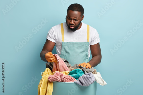 Puzzled Afro American man overstained with housekeeping, does laundry at home during Saturday, looks frustratedly at towel, notices stain, dissatisfied with wash, wears t shirt and blue apron photo