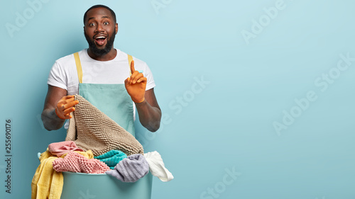 Satisfied black man raises fore finger as gets brilliant idea how to wash off stains on rag, poses near basin full of dirty linen, dressed in casual clothes, isolated over blue wall with free space photo