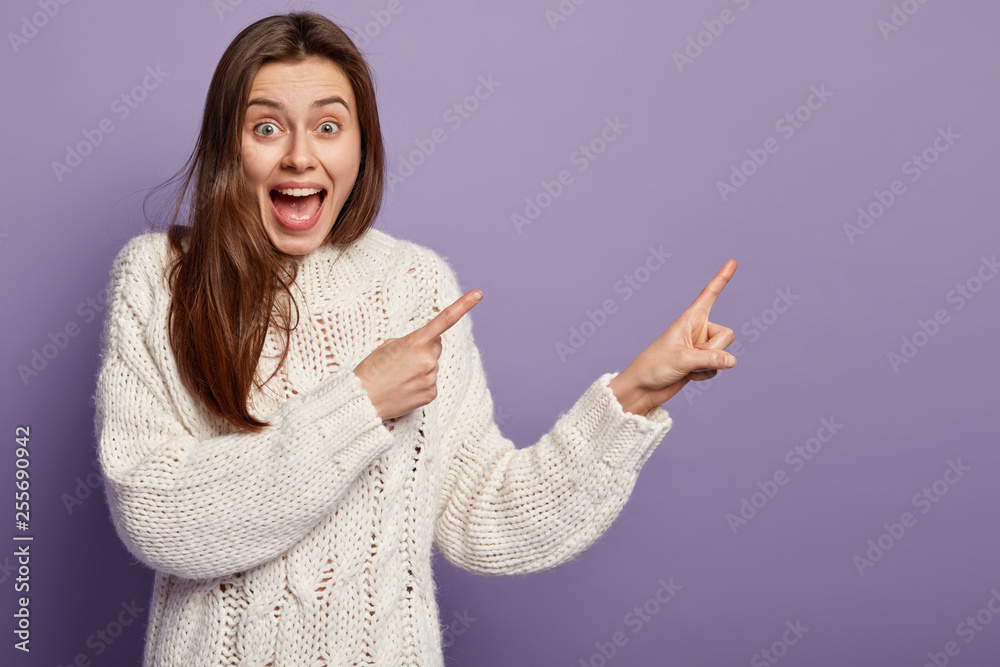 Photo of happy European woman points away with both fore fingers, glad to advertise something, indicates index fingers aside, wears white clothes, isolated over purple background. Promotion concept Photo of happy European woman points away with both fore fingers, glad to advertise something, indicates index fingers aside, wears white clothes, isolated over purple background. Promotion concept