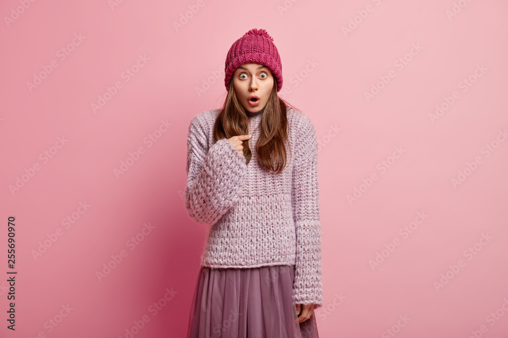 Bugged eyed European woman points at herself, stares in surprisement, indicates at herself, asks why she should do this, wears headgear, loose jumper and skirt, isolated over pink studio wall Bugged eyed European woman points at herself, stares in surprisement, indicates at herself, asks why she should do this, wears headgear, loose jumper and skirt, isolated over pink studio wall