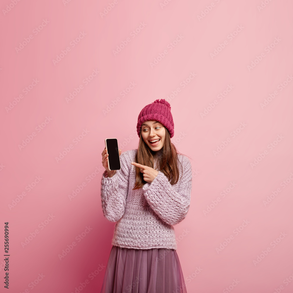 Vertical shot of pleased European woman in pink headgear, oversized jumper and pleated skirt points at cell phone, shows mock up black screen for your advertisement, stands over pink background Vertical shot of pleased European woman in pink headgear, oversized jumper and pleated skirt points at cell phone, shows mock up black screen for your advertisement, stands over pink background