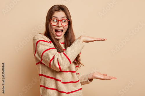 Photo of cheerful woman boasts of present size she received, shapes big object with both hands, demonstrates height of thing, wears striped sweater, optical glasses, stands over beige background photo