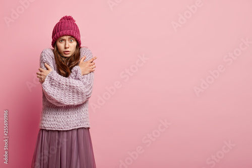 Omg, so cold. Lovely young female crosses hands over chest, wears knitted sweater, headgear, has walk during cold winter day, models over pink background with free space, clatters with teeth