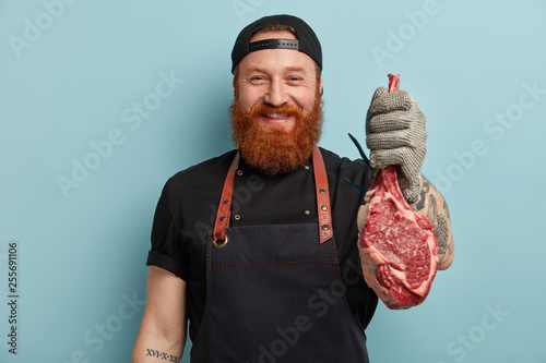Glad cheerful butcher suggests to taste beef or pork, works on meat factory, cuts on small pieces for selling, wears special uniform, isolated over blue background. Food industry and processing
