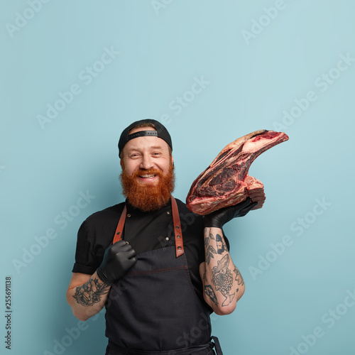 Meat consumption concept. Satisfied man meatpaking worker, smiles gladfully, carries raw pork on bone, works on slaughthouse, keeps hand on apron, wears protective gloves, isolated on blue wall photo