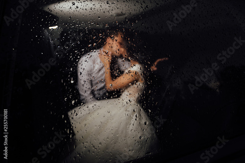 The bride and groom kissing in the car late at night, and it's raining outside. Bride and groom photographed behind the wet glass