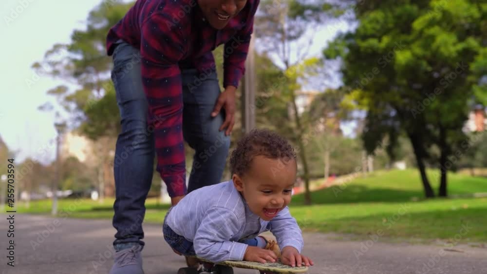 Little mixed-race boy in park wearing blue shirt, jeans lying on skateboard with face down, rolling with help of Afro-american young father in striped shirt and jeans. Family, game concept Little mixed-race boy in park wearing blue shirt, jeans lying on skateboard with face down, rolling with help of Afro-american young father in striped shirt and jeans. Family, game concept