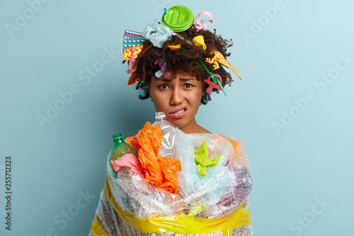 Plastic pollution problem. Dissatisfied woman wrapped in polythene sheeting, collected much garbage, frowns from dislike, isolated over blue background. Save Earth concept. Clean our nature. photo