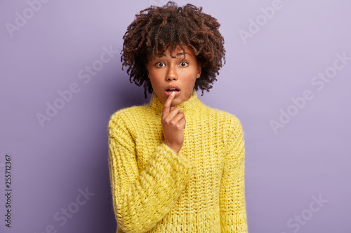 Confused focusd young woman keeps hand near mouth  expresses wonder as listens something surprising  wears yellow jumper  has dark healthy skin  Afro hairstyle  isolated on purple background