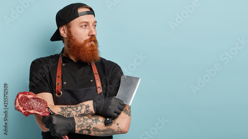 Sideways shot of handsome thoughtful male meat cutter prepares cuts of meat for selling, involved in idustrial processing, carries ceaver and raw meat, stands over blue background with free space photo