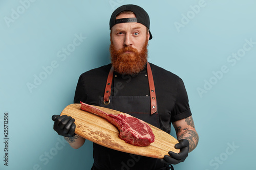 Horizontal shot of self confident satisfied bearded young male cook looks with serious expression, wears cap and black apron, holds cutting board with raw meat, isolated over blue background photo