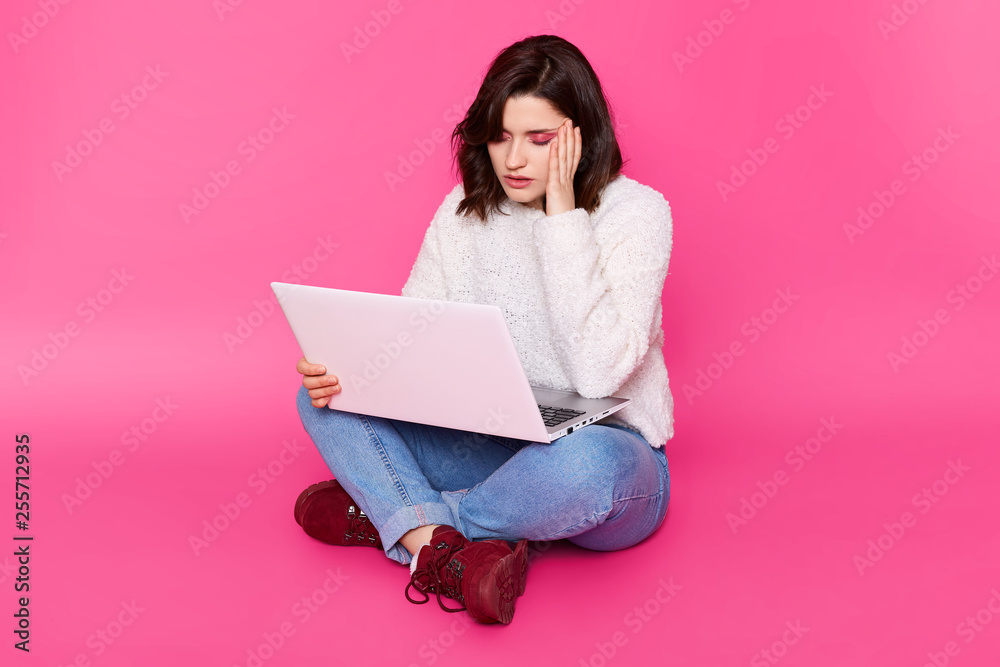 Tired student with laptop sits on floor with closed eyes. Brunette girl keeps hand near temple because sufers from headache. Female works online during long hours. Model poses against pink wall. Tired student with laptop sits on floor with closed eyes. Brunette girl keeps hand near temple because sufers from headache. Female works online during long hours. Model poses against pink wall.