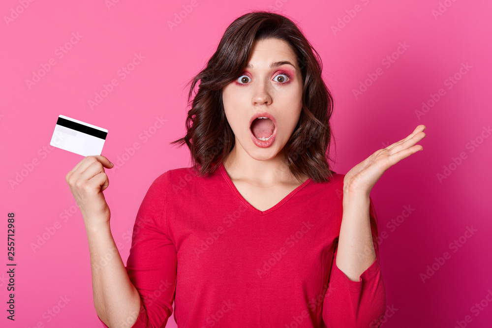 Astonished brunette woman being short of money, has no opportunity to make purchase online. Young female wears sewater stands with opened mouth isolated on pink wall. Interent and shopping concept. Astonished brunette woman being short of money, has no opportunity to make purchase online. Young female wears sewater stands with opened mouth isolated on pink wall. Interent and shopping concept.