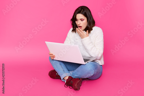 Amazed beautiful woman looks on laptop screen with wide opened eyes, sits with crossed legs on floor, keeps hand on mouth. Girl sees shocking best friends photos with her boyfriend in social network. © sementsova321